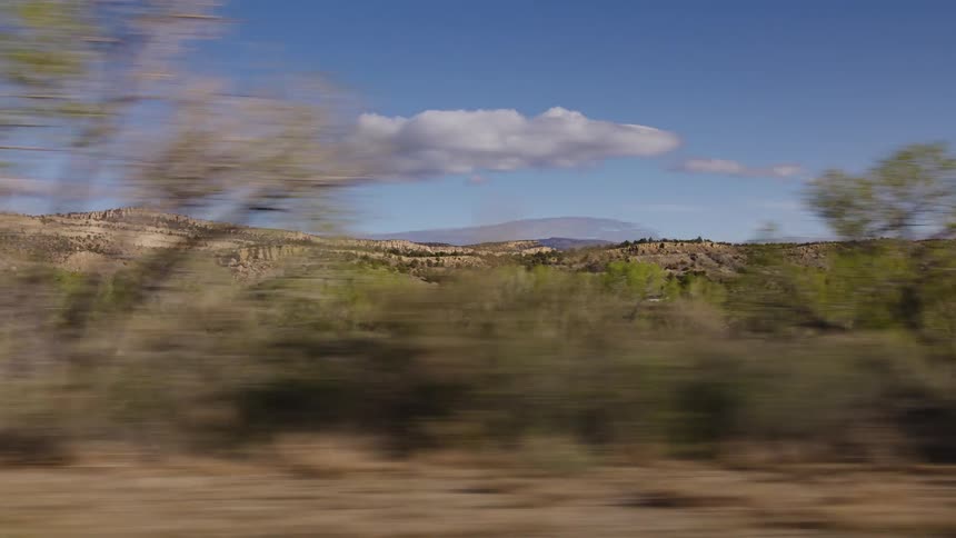 A scenic drive through a desert landscape reveals rocky hills and green trees under a blue sky, captured from a moving vehicle