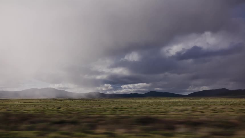 A passenger's view of a vast, rolling prairie under a brooding, stormy sky with rain falling on distant mountains