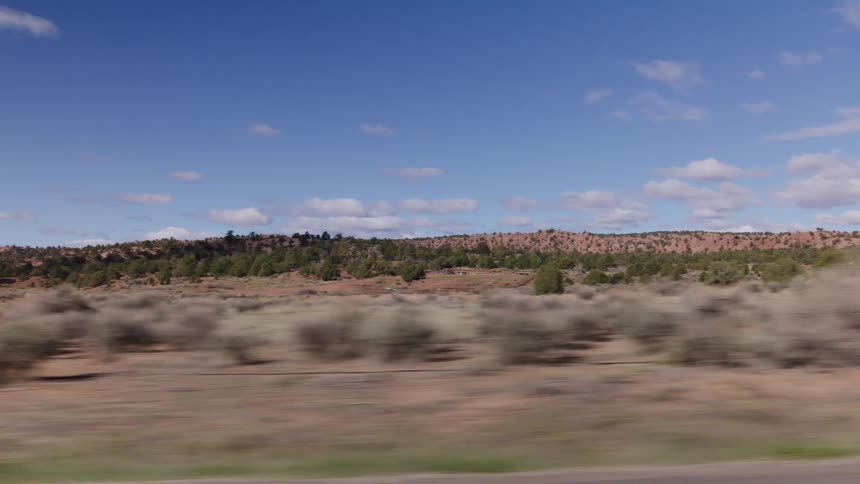 A passenger's view from a moving car reveals a vast desert landscape with scrub brush and distant hills under a blue sky