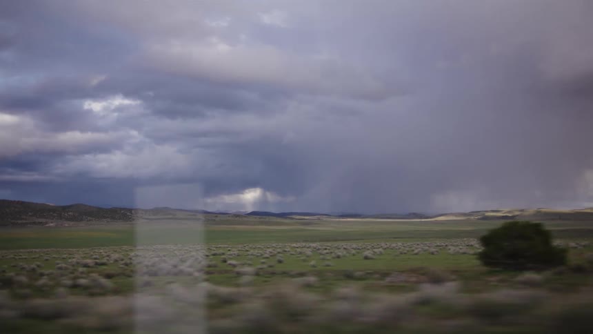 A slow-motion tracking shot from a vehicle captures a dramatic rainstorm over a vast, open green landscape