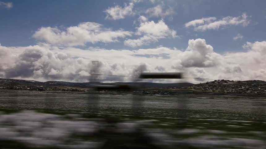 A scenic view from a moving vehicle captures a snow-dusted landscape with a tranquil lake and a distant town under a dramatic sky