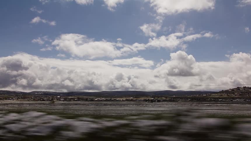 A scenic side-scrolling view from a moving vehicle of a vast, snowy landscape with a distant town under a cloudy blue sky