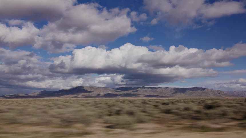 Side view from a moving vehicle captures the sweeping desert landscape and mountain range under a partly cloudy sky