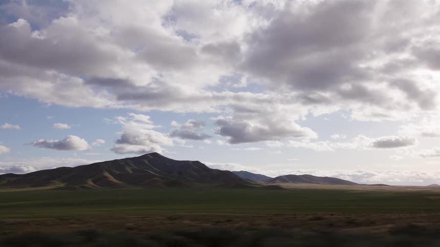 A scenic view of a vast green valley and rolling hills under a cloudy sky as seen from a moving vehicle