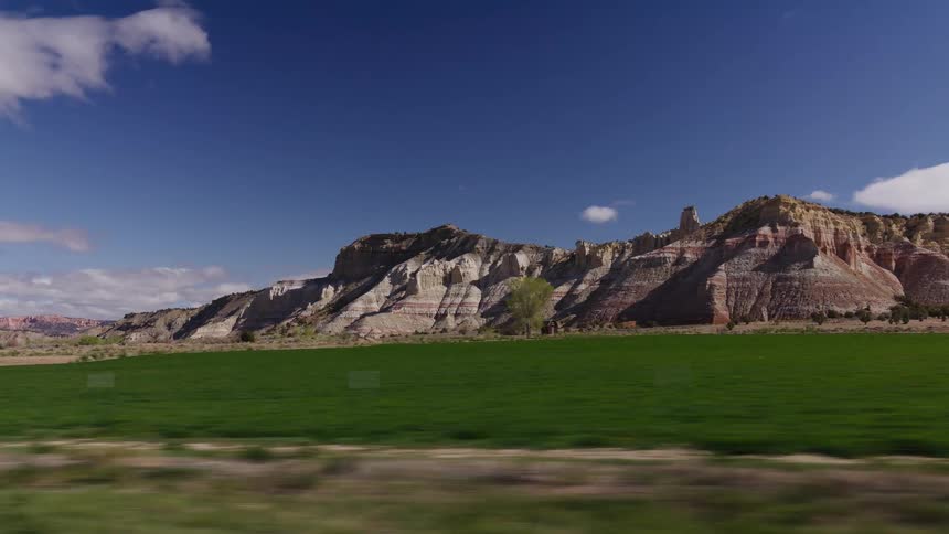 A scenic drive past colorful, stratified rock formations and pastoral fields under a clear blue sky in the American Southwest