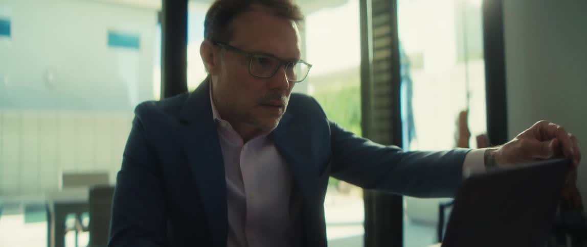 A focused middle-aged businessman in a suit works intently on his laptop, then takes off his glasses