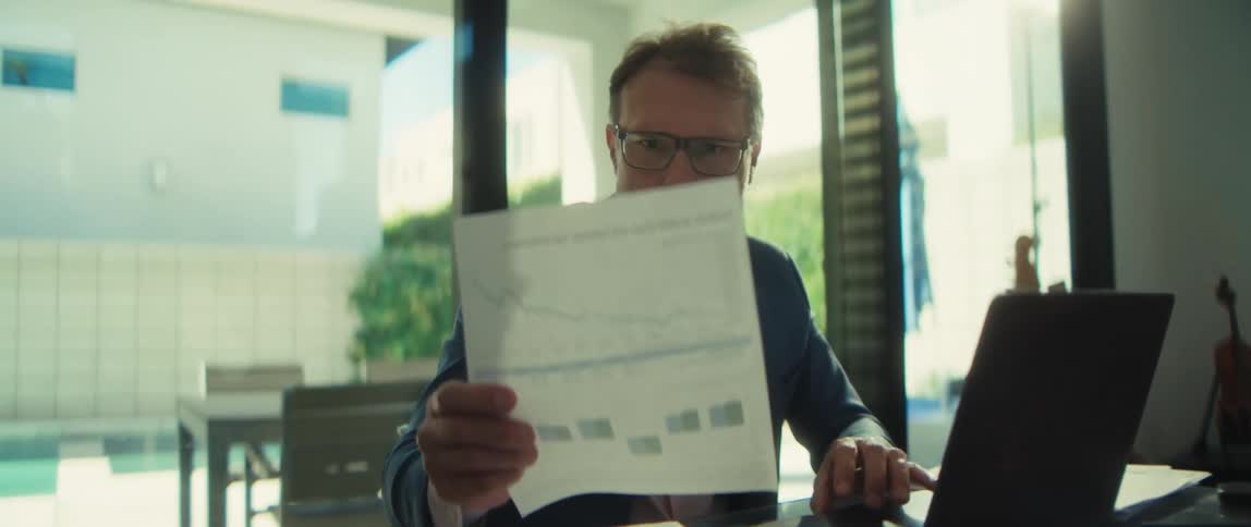 A focused professional man in a blazer analyzes financial documents and works on his laptop in a bright, contemporary home office