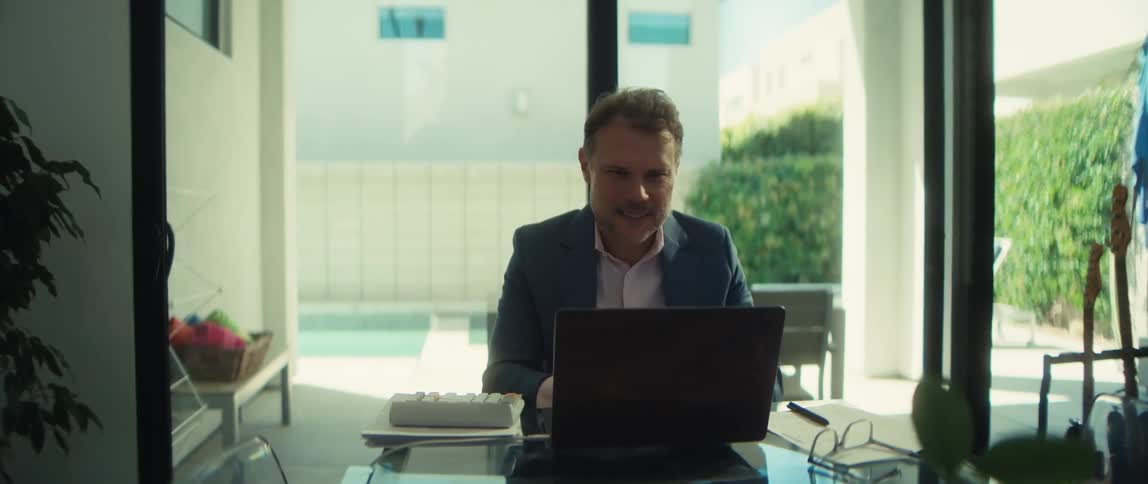 A happy businessman in a suit jacket works remotely from his modern home office, smiling as he types on his laptop