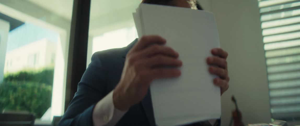 A focused businessman in a suit diligently reviews important documents before turning to work on his laptop in a sunlit modern office