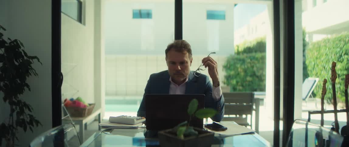 A stressed businessman in his home office holds his glasses and rubs his temple as he works on his laptop