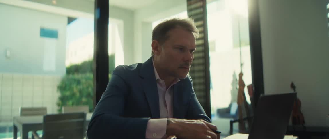 A slow push-in shot reveals a serious businessman working on his laptop and taking notes in his sunlit modern home