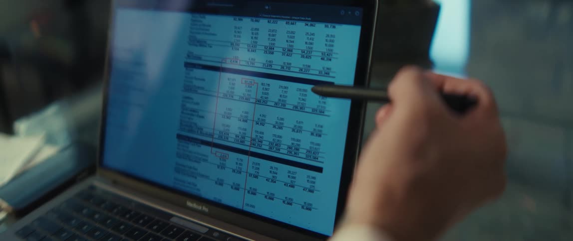 A financial analyst uses a stylus pen to review a spreadsheet with columns of data on a laptop screen