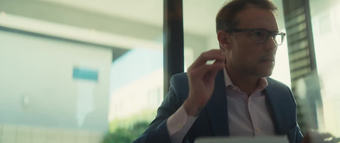 A professional businessman in a suit jacket puts on his eyeglasses to focus on his work in a modern, sunlit office