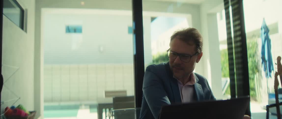 A professional man in a suit jacket works diligently from his modern home office, with a sunny pool visible in the background