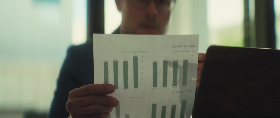 A focused businessman in glasses meticulously examines financial bar graphs on a document while working at his desk in a modern office