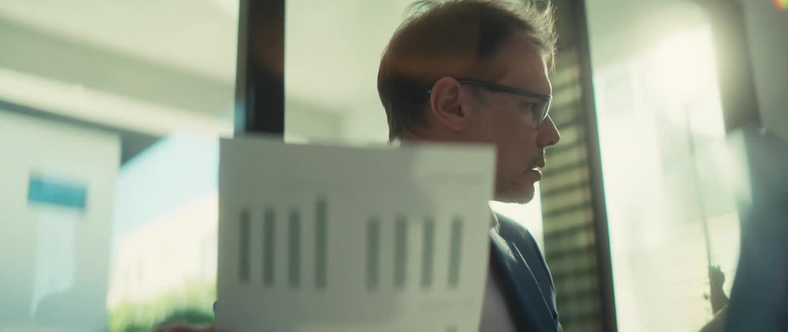 A close-up of a businessman in glasses carefully analyzing a business report with bar charts in a brightly lit office