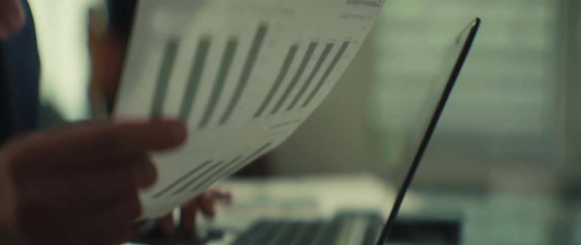 A businessman in glasses analyzes a financial bar graph report while working on his laptop in a warmly lit office