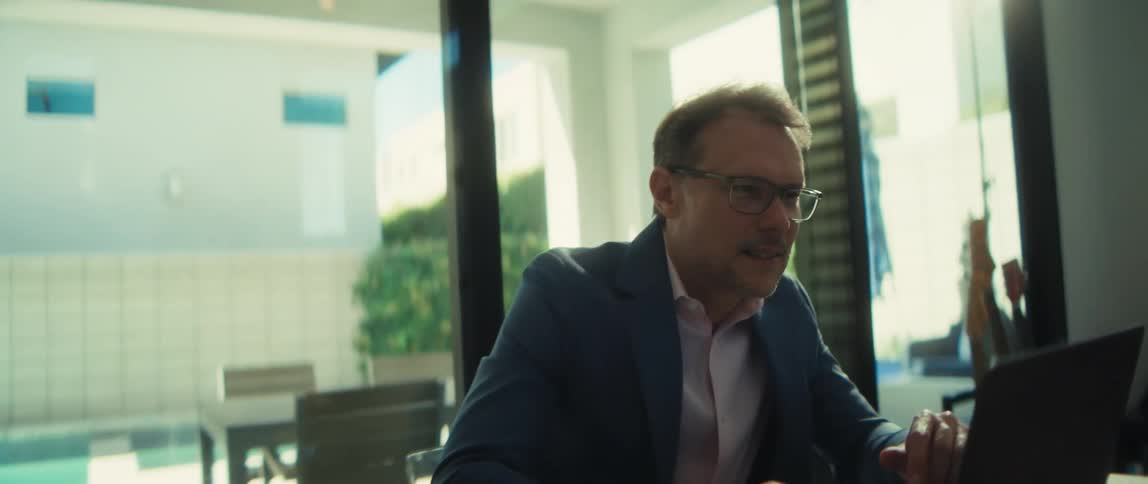 Dolly-in shot of a smiling middle-aged businessman working remotely on a laptop in his bright, modern home