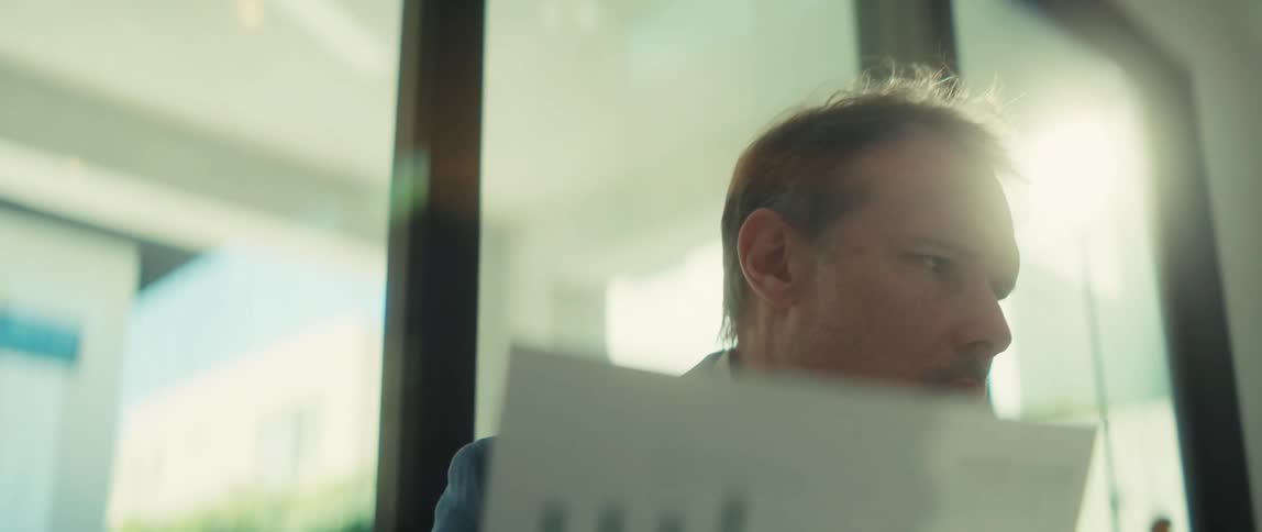 A concerned businessman in a modern, sun-drenched office removes his glasses while analyzing a financial report