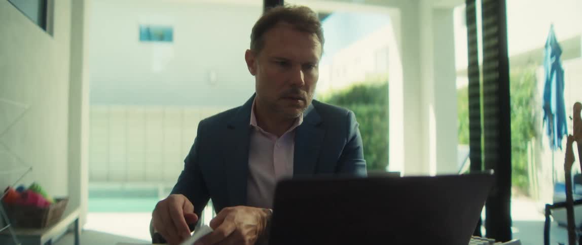 A focused businessman diligently works from his modern home office, taking off his glasses while reviewing documents and using his laptop