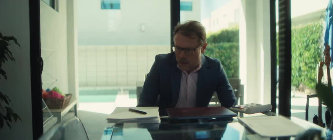 A middle-aged businessman in a suit walks to his desk in a modern home office, sits down, and opens his laptop to begin working