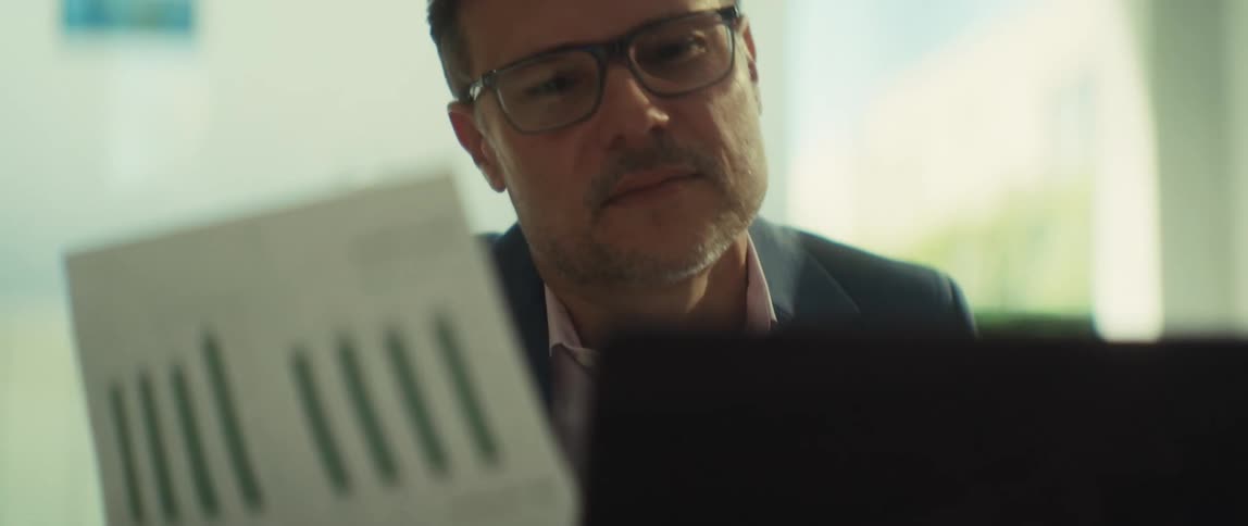 A focused businessman in glasses meticulously compares a printed bar chart with data on his laptop while working in a bright, modern office