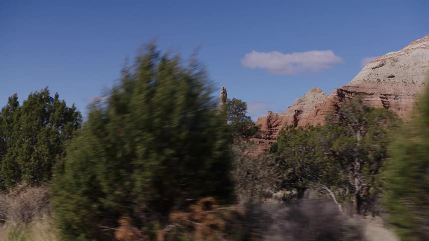 A slow-motion driving shot reveals a majestic red rock spire and layered cliffs in a desert landscape under a clear blue sky