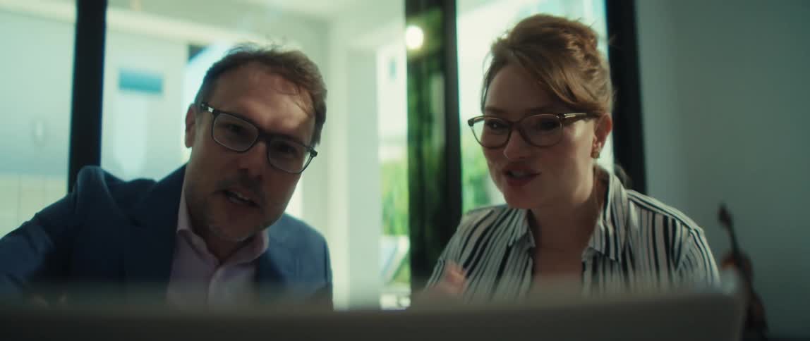 A professional man and woman in glasses collaborate and smile while looking at a laptop screen together in a modern office