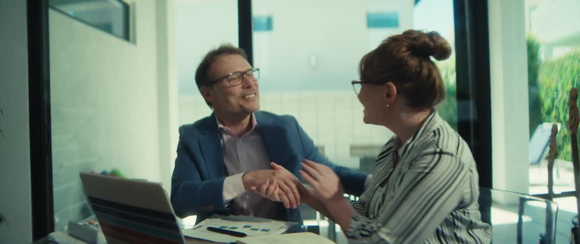 In slow motion, a businessman and businesswoman smile and shake hands, finalizing a successful deal in a contemporary office space