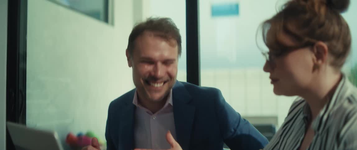 A smiling businessman claps his hands in celebration after his female colleague presents successful results on a laptop in their office