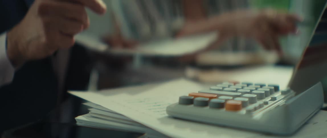 A businessman in a suit uses a calculator at his desk while reviewing financial paperwork in his office