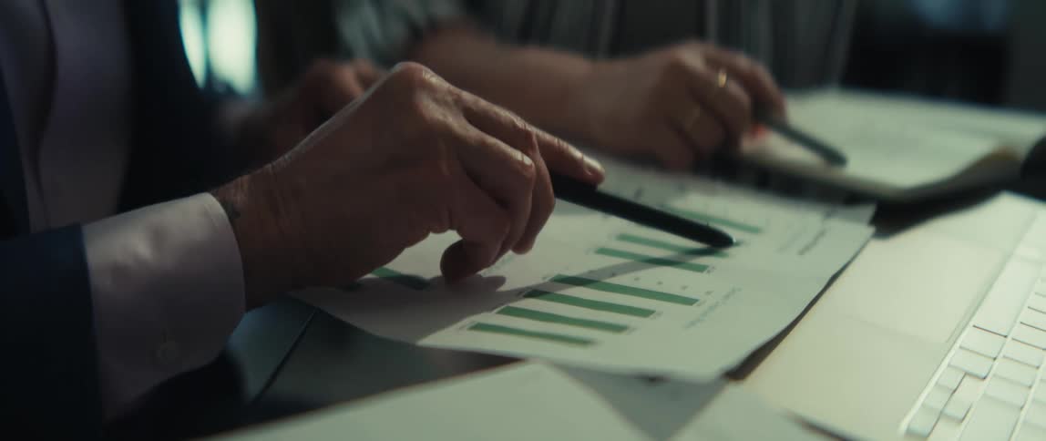 Close-up of two colleagues' hands pointing with pens to analyze business data on a green bar chart document