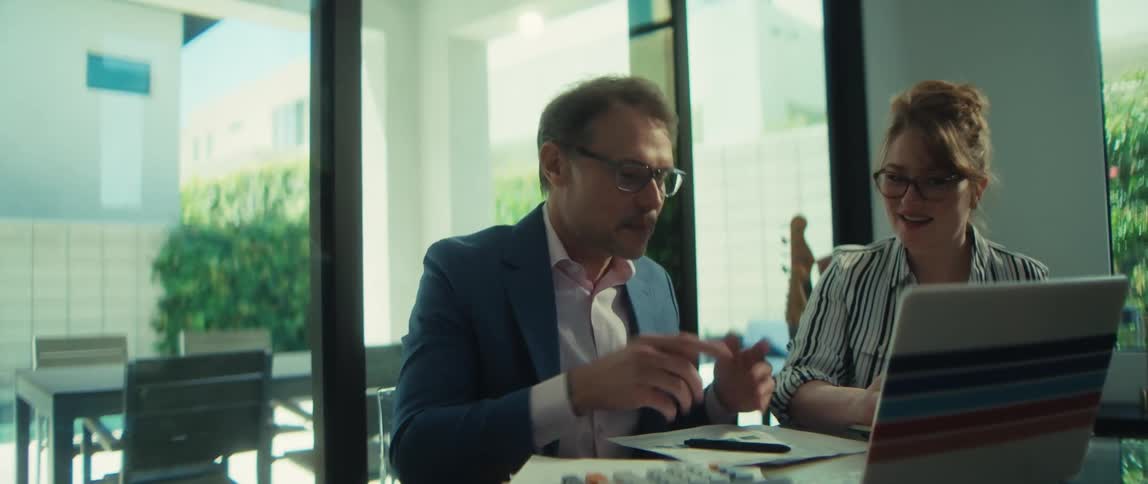 A male and female colleague discuss a project on a laptop while sitting at a glass table in a modern, naturally lit office