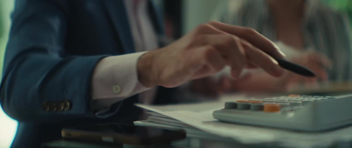 A businessman in a suit explains financial documents to a female colleague while using a calculator in a modern office