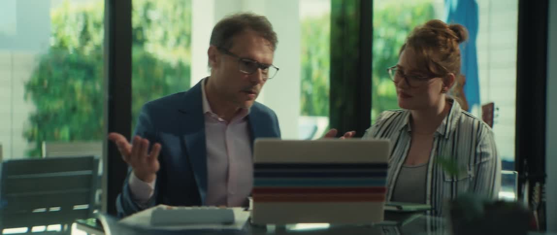 A professional man and woman wearing glasses collaborate on a project, having an engaged discussion over a laptop in a modern, sunlit office
