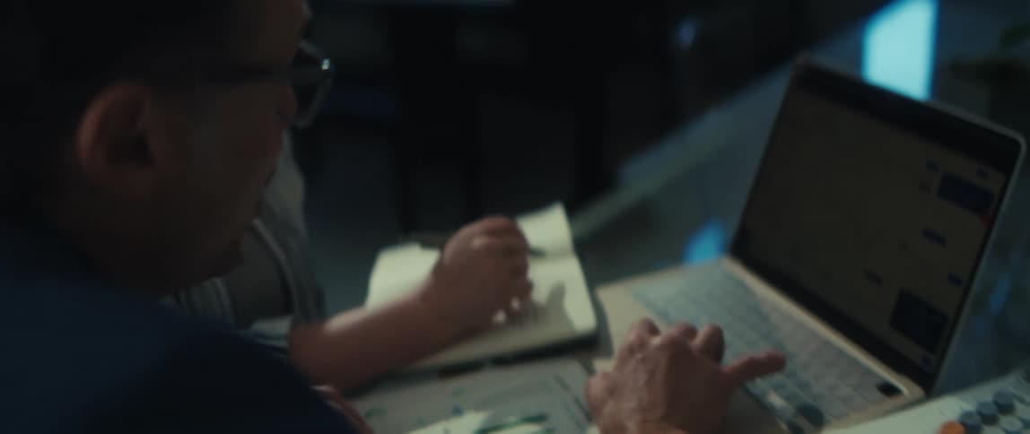 Two financial analysts collaborate over a laptop, examining stock market charts while one takes notes in a notebook