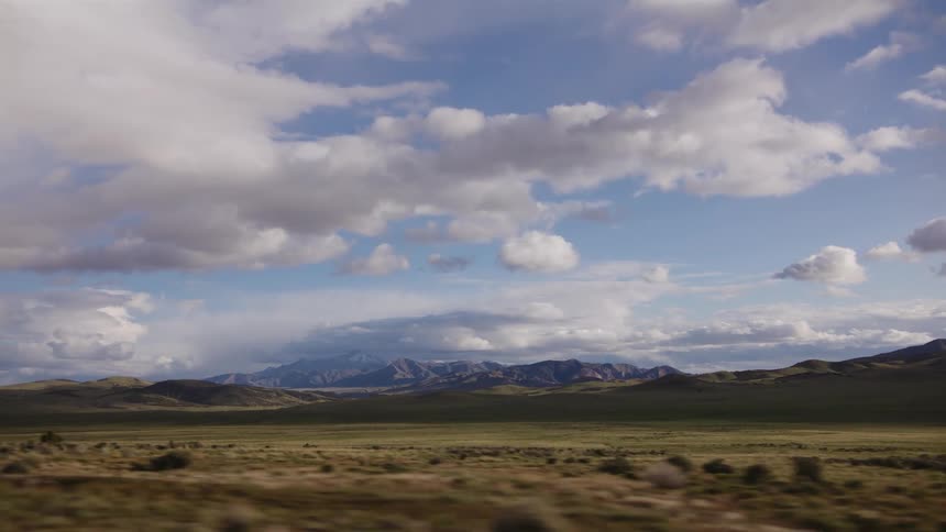 A scenic drive-by view of a vast desert valley with a majestic mountain range under a cloudy blue sky