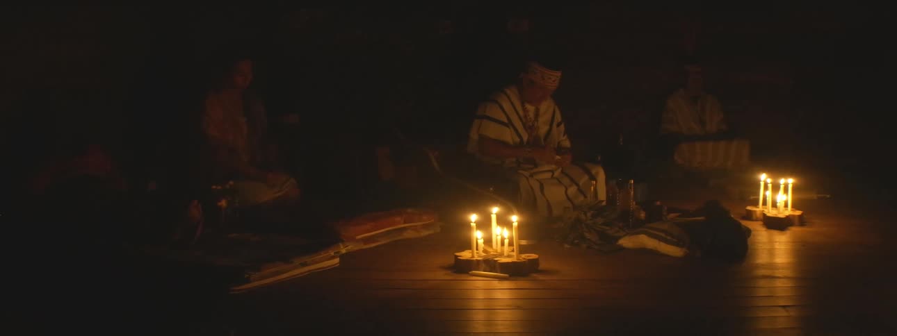 An indigenous shaman leads a traditional spiritual ceremony by candlelight in a dark, sacred space