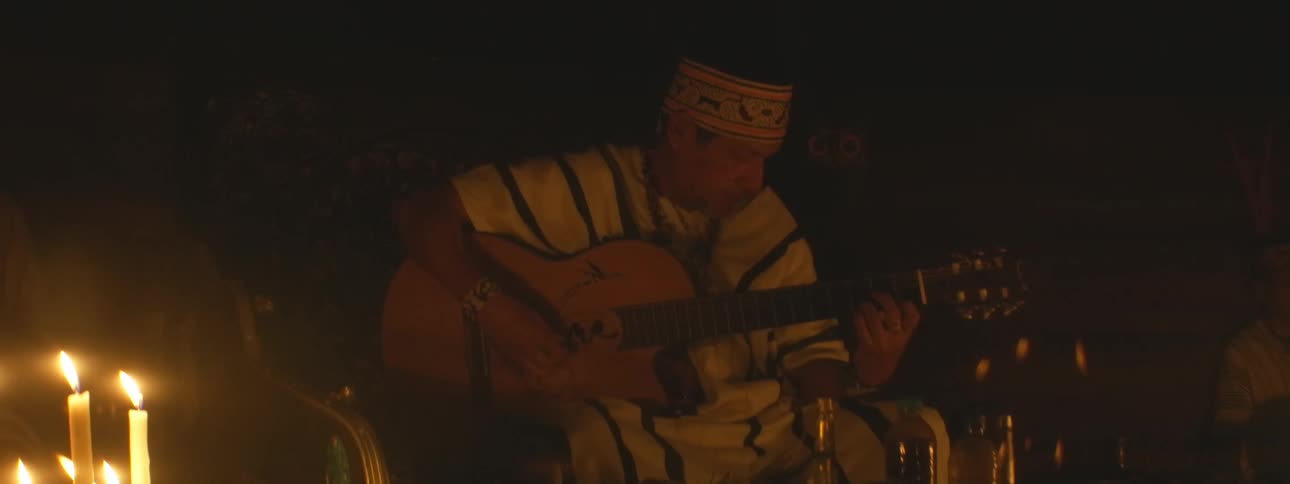 An indigenous musician in traditional dress plays a soulful melody on an acoustic guitar in a dark, candlelit setting