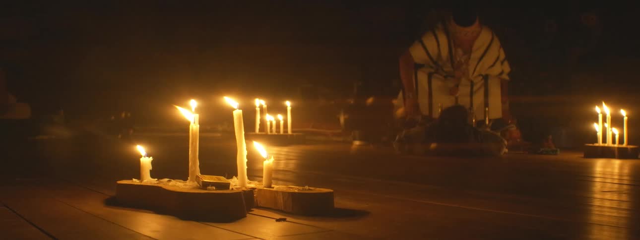 A low-angle shot captures flickering candles illuminating a traditional shamanic ceremony in a dark, sacred space