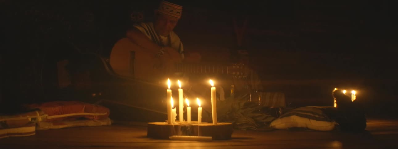 An indigenous shaman leads a spiritual ceremony by candlelight, playing a gentle song on his acoustic guitar