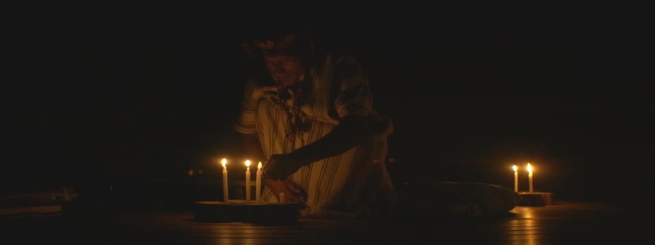 A shaman in traditional attire kneels in the dark, methodically lighting candles in preparation for a sacred spiritual ceremony