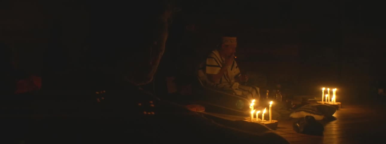 An indigenous shaman leads a spiritual healing ceremony, chanting and smoking in a dark room illuminated only by flickering candles