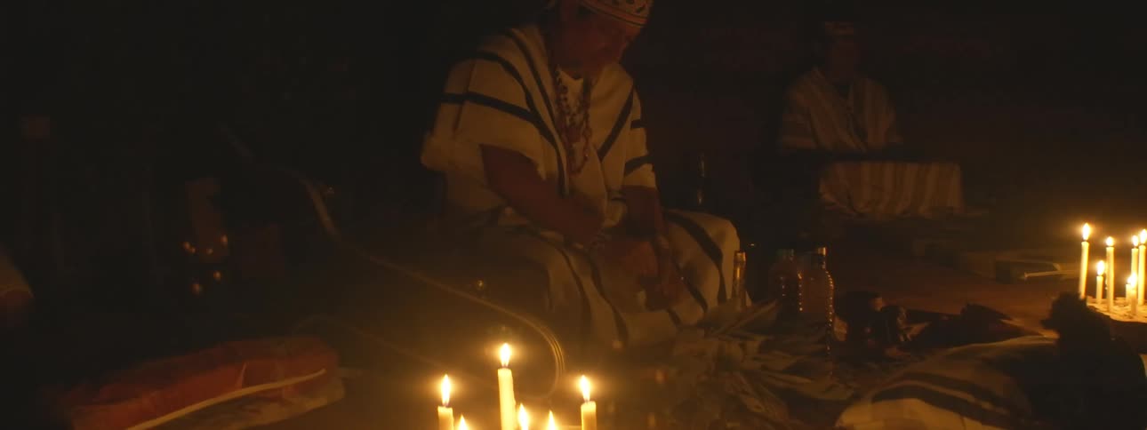 An indigenous shaman in traditional clothing performs a sacred spiritual ritual by the warm glow of candlelight at night