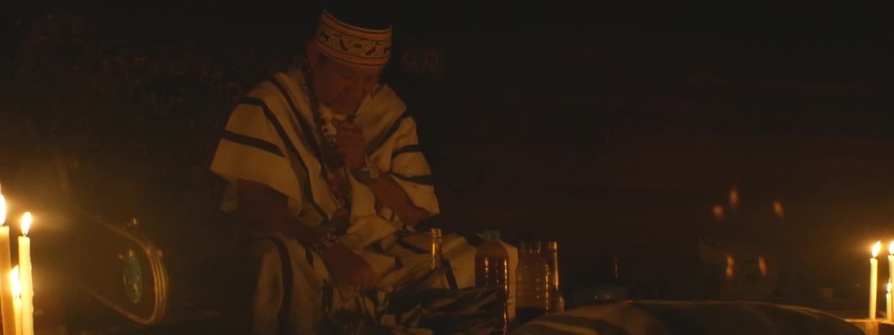 An indigenous shaman in traditional attire meditates during a spiritual ceremony in a dimly lit, candlelit room