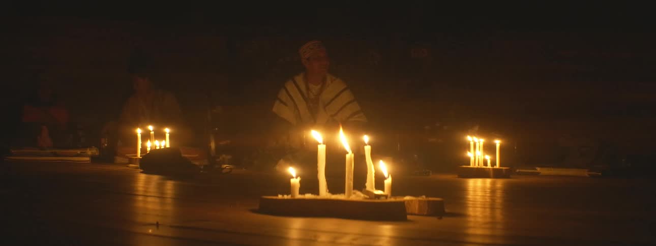A shaman leads a spiritual ceremony in a dark room illuminated only by the flickering light of many candles