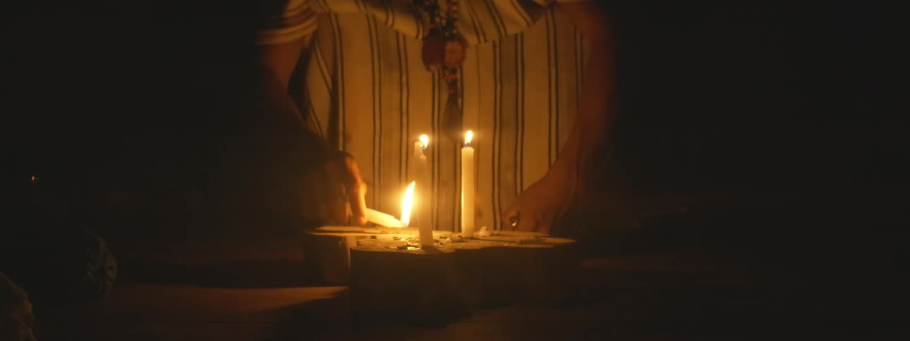 An indigenous shaman in traditional attire lights several candles in slow motion for a spiritual ceremony in a dark room