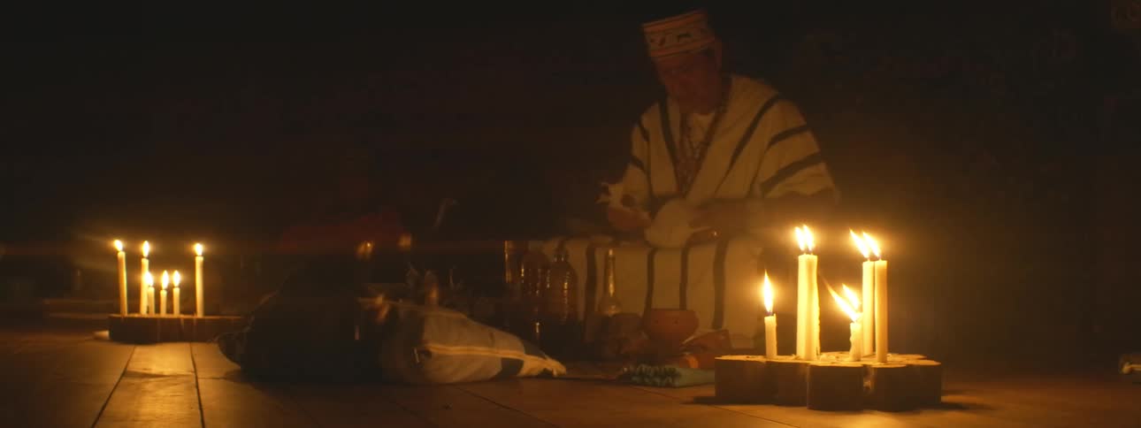 An indigenous shaman in traditional clothing leads a spiritual healing ceremony at night surrounded by flickering candles