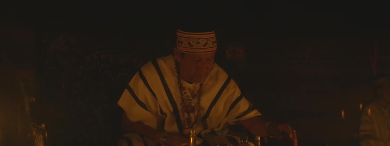 A shaman in traditional garments performs a spiritual cleansing ritual at night, smoking tobacco and waving a bundle of sacred leaves