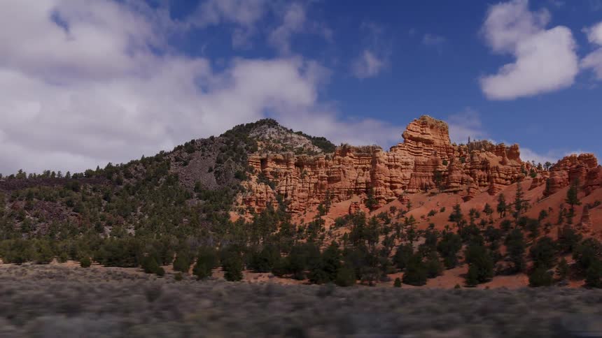 A scenic tracking shot reveals the breathtaking red rock cliffs and forested hillsides of Southern Utah under a partly cloudy sky
